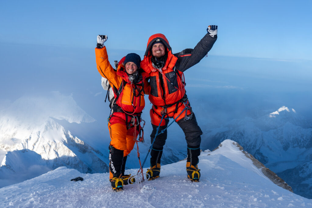 Man and woman standing top of mountain with background of more of mountains.